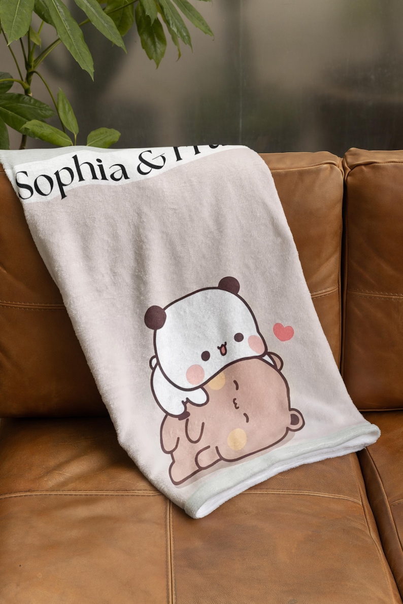 May include: A soft, light pink throw blanket with a white border. The blanket features a cute cartoon panda bear hugging a brown bear. The text "Sophia &" is printed on the top of the blanket.