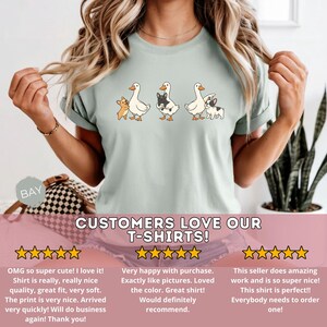 May include: Light green t-shirt featuring a whimsical design of three geese with small dog companions. The geese are white with orange beaks and feet, and the dogs are in various colors. The text "CUSTOMERS LOVE OUR T-SHIRTS!" is displayed.