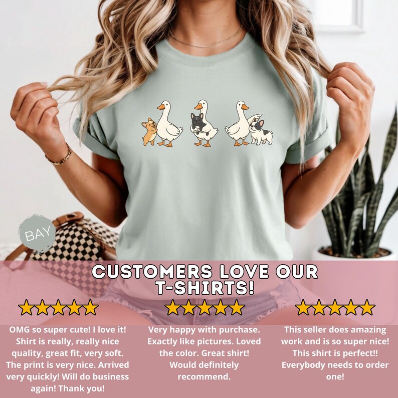 May include: Light green t-shirt featuring a whimsical design of three geese with small dog companions. The geese are white with orange beaks and feet, and the dogs are in various colors. The text "CUSTOMERS LOVE OUR T-SHIRTS!" is displayed.