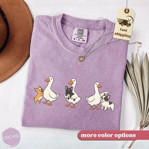 May include: A lilac-colored t-shirt with a crew neck featuring a graphic of three white ducks with orange beaks and feet, each accompanied by a small dog. The shirt has a Comfort Colors tag and a silver necklace. A tag reads "fast shipping".