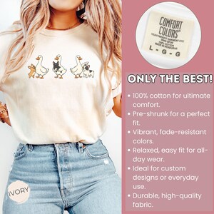 May include: Ivory-colored t-shirt featuring a whimsical design of geese and dogs. The shirt has a crew neck and short sleeves. The text "COMFORT COLORS" is visible on a tag. The shirt is made of 100% cotton.
