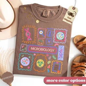 May include: Espresso-colored t-shirt with a microbiology design. The shirt features colorful illustrations of cells, DNA, and lab tools. The word "MICROBIOLOGY" is displayed in a rectangular frame. A gold necklace and sandals are also visible.