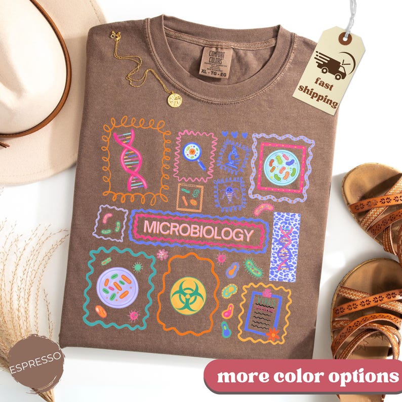 May include: Espresso-colored t-shirt with a microbiology design. The shirt features colorful illustrations of cells, DNA, and lab tools. The word "MICROBIOLOGY" is displayed in a rectangular frame. A gold necklace and sandals are also visible.