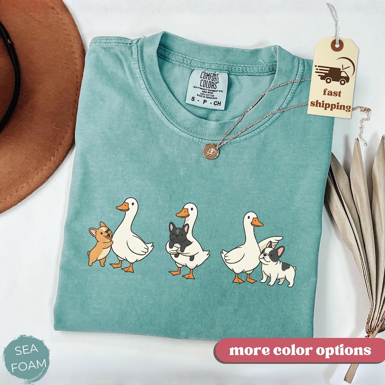 May include: Seafoam green Comfort Colors t-shirt with a graphic of three white ducks with orange beaks and feet, each paired with a small dog. The shirt has a crew neck and short sleeves. A gold necklace and a tag that says "Fast Shipping" are also visible.