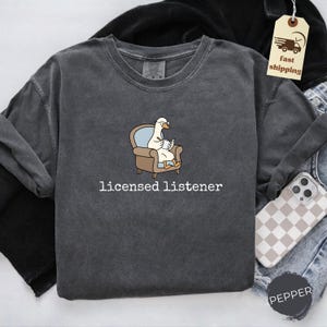 May include: Dark gray long-sleeve shirt with the text "licensed listener." The shirt features a cartoon goose wearing glasses and reading a book while seated in a chair. The shirt is made of a soft, comfortable material.