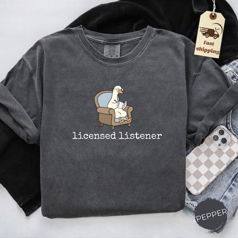 May include: Dark gray long-sleeve shirt with the text "licensed listener." The shirt features a cartoon goose wearing glasses and reading a book while seated in a chair. The shirt is made of a soft, comfortable material.