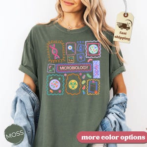 May include: Moss green t-shirt with a colorful microbiology design. The design features various scientific illustrations and the word "MICROBIOLOGY" in a rectangular box. The shirt has rolled-up sleeves and a relaxed fit.