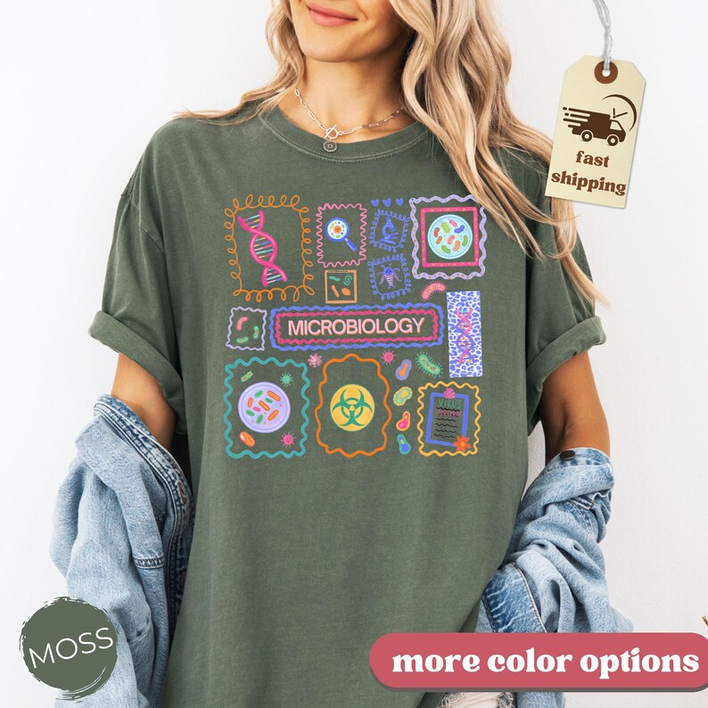 May include: Moss green t-shirt with a colorful microbiology design. The design features various scientific illustrations and the word "MICROBIOLOGY" in a rectangular box. The shirt has rolled-up sleeves and a relaxed fit.