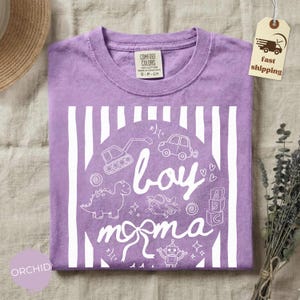 May include: Orchid-colored t-shirt with a white graphic that says "boy moma" with illustrations of a dinosaur, car, airplane, robot, and other toys. The shirt has a crew neck and is made of a soft material.
