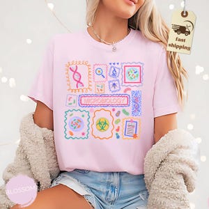 May include: A light pink t-shirt featuring a colorful graphic design related to microbiology. The design includes various scientific illustrations within decorative frames, with the word "MICROBIOLOGY" prominently displayed. The shirt is made of a soft material.