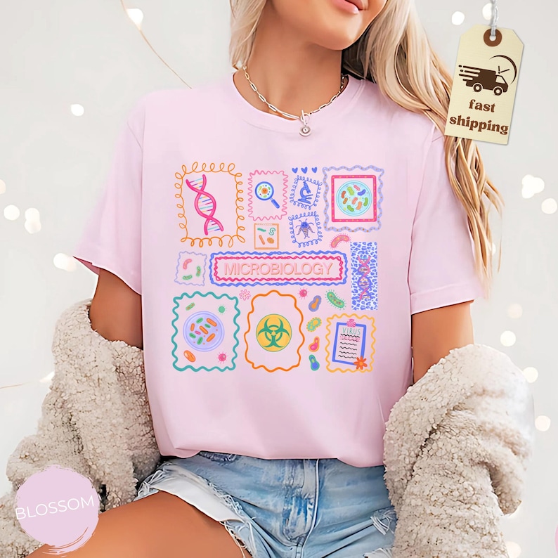 May include: A light pink t-shirt featuring a colorful graphic design related to microbiology. The design includes various scientific illustrations within decorative frames, with the word "MICROBIOLOGY" prominently displayed. The shirt is made of a soft material.