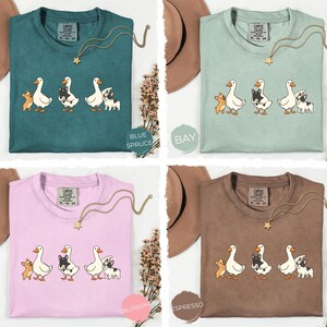 May include: Four Comfort Colors t-shirts in blue spruce, bay, blossom, and espresso. Each shirt features a graphic of three animals: a duck, a black and white dog, and a tan dog. Each shirt has a gold necklace.
