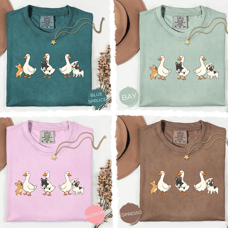 May include: Four Comfort Colors t-shirts in blue spruce, bay, blossom, and espresso. Each shirt features a graphic of three animals: a duck, a black and white dog, and a tan dog. Each shirt has a gold necklace.