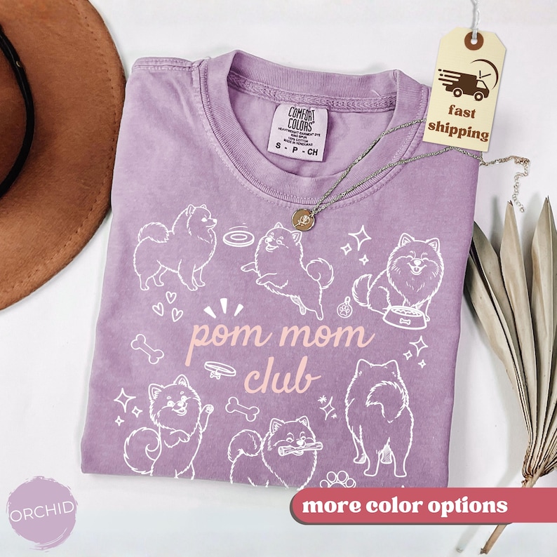 以下が含まれることがあります： クルーネックのライラック色のTシャツ。白いポメラニアン犬のグラフィックと「pom mom club」の文字が特徴です。ゆったりとしたフィット感で、「Comfort Colors」のタグが付いています。ペンダント付きのシルバーネックレスがシャツの上にかけられています。