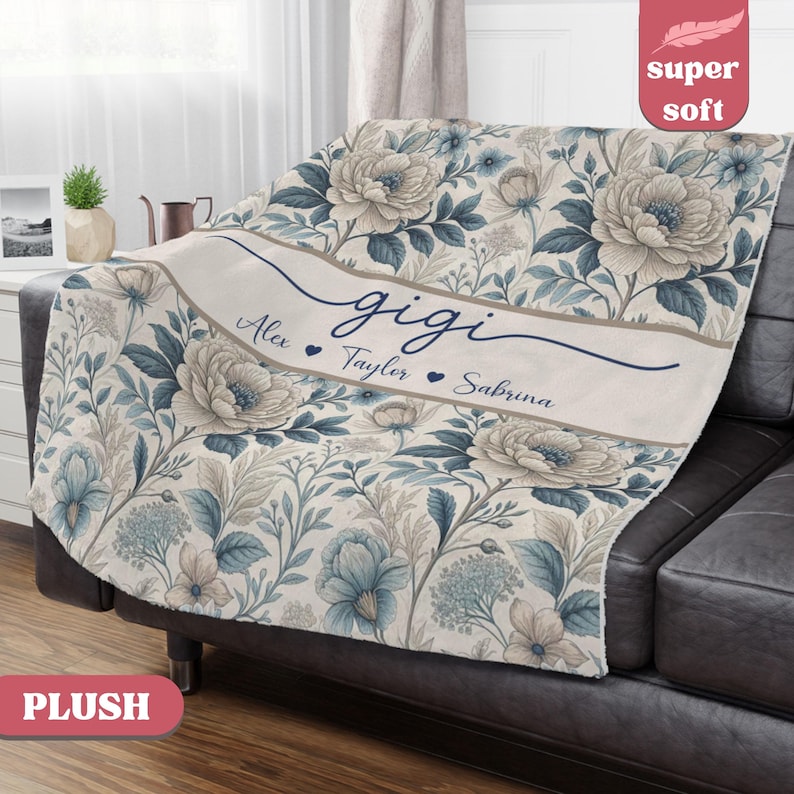 Puede incluir: Una manta de felpa con un estampado floral en tonos azules, crema y grises. La manta presenta el nombre "gigi" en cursiva, con nombres debajo. La manta est&aacute; sobre un sof&aacute; gris oscuro.