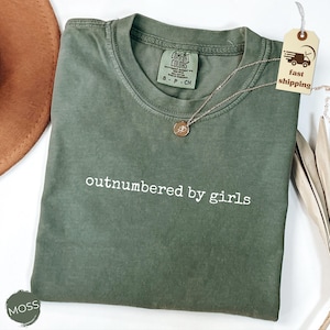 May include: An olive green t-shirt with the phrase "outnumbered by girls" in white lettering. A gold necklace with a small pendant rests on the shirt. A tag with a truck graphic and the words "fast shipping" is attached.