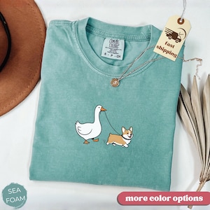 May include: Seafoam green Comfort Colors t-shirt featuring a cartoon goose walking a corgi on a leash. The shirt has a crew neck and short sleeves. A tag with "fast shipping" is attached. The words "more color options" are at the bottom.
