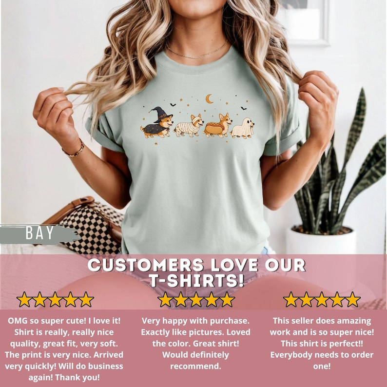 May include: A light sage green short-sleeved t-shirt featuring a graphic print of four Corgi dogs dressed in Halloween costumes, including a witch, a mummy, a hot dog, and a ghost, accented with small stars, a crescent moon, and bats.