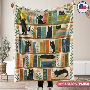 May include: A cream-coloured sherpa fleece blanket with a black cat and book illustration. The illustration features a bookshelf with black cats reading books, sleeping on books, and sitting on books. The bookshelf is decorated with green leaves and flowers.
