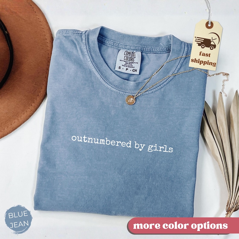 May include: A blue jean colored t-shirt with the text "outnumbered by girls" in white. The shirt has a crew neck and a small tag on the collar. A gold necklace and a brown hat are also in the image.
