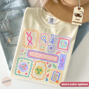 May include: Ivory-colored sweatshirt featuring a colorful microbiology-themed design. The design includes various scientific illustrations, such as DNA strands, microscopes, and bacteria, framed in decorative borders. The word "MICROBIOLOGY" is prominently displayed.