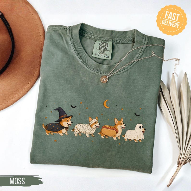 May include: A moss green crewneck t-shirt featuring a graphic of four Corgi dogs in Halloween costumes, including a witch, a mummy, a hot dog, and a ghost, with small stars, a crescent moon, and bats printed above the dogs on the chest area.