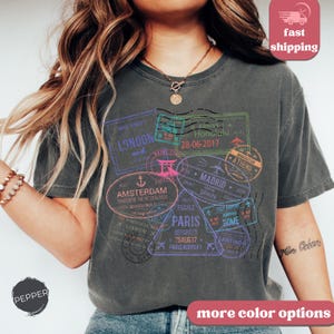 May include: Dark gray t-shirt featuring travel-themed stamps with city names like London, Paris, and Amsterdam. The stamps are in various colors, including purple, green, and pink. The shirt has the text "PEPPER" and "more color options".