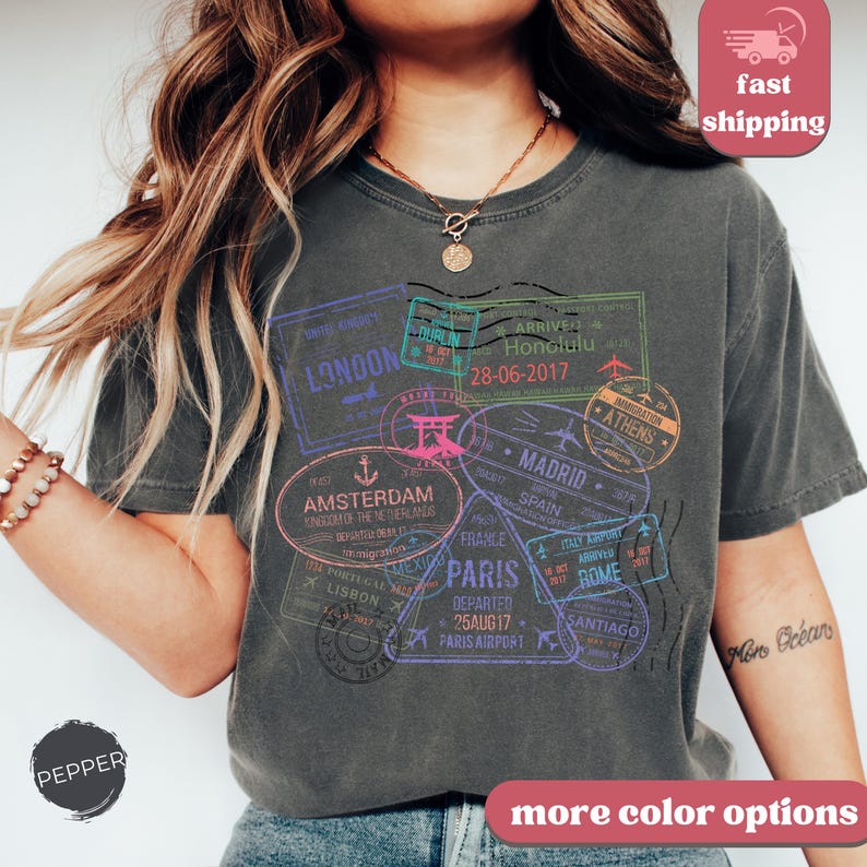 May include: Dark gray t-shirt featuring travel-themed stamps with city names like London, Paris, and Amsterdam. The stamps are in various colors, including purple, green, and pink. The shirt has the text "PEPPER" and "more color options".