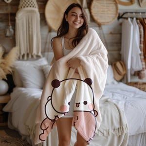 May include: A white plush blanket with a panda bear design. The panda is smiling and has a pink nose and tongue. The blanket is being held by a person.