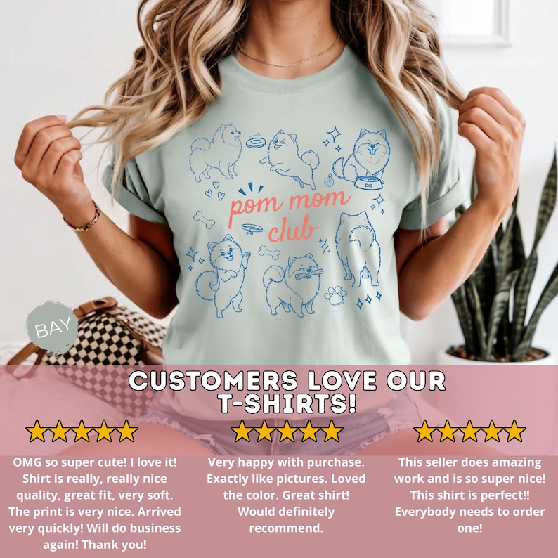 以下が含まれることがあります： 水色のTシャツ。青いポメラニアン犬のデザインとピンクの文字「pom mom club」が特徴です。Tシャツには「CUSTOMERS LOVE OUR T-SHIRTS！」の文字も入っています。デザインには骨と足跡が含まれています。