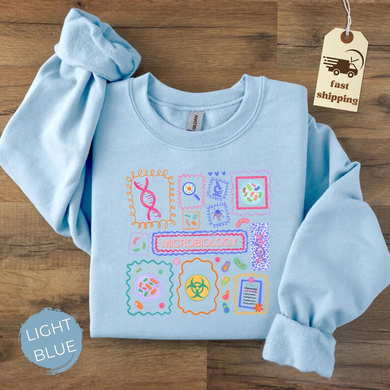 May include: Light blue sweatshirt with a colorful microbiology-themed graphic. The design includes various scientific illustrations and the word "MICROBIOLOGY" in a rectangular frame. The sweatshirt has long sleeves and a crew neck.