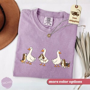 May include: A lilac-colored t-shirt featuring a graphic of three white geese with orange beaks and feet, each accompanied by a brown and white basset hound. The shirt has a crew neck and short sleeves. A tag with "fast shipping" hangs from the shirt.