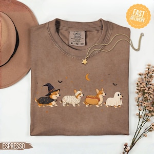 May include: A brown crewneck t-shirt featuring a graphic of four Corgi dogs in Halloween costumes: a witch, a mummy, a hot dog, and a ghost. The shirt has a Comfort Colors label, a crescent moon, stars, and bats printed above the dogs.