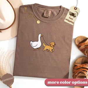 Pu&ograve; includere: T-shirt color espresso con un cartone animato di un'oca che porta a spasso un golden retriever al guinzaglio. La maglietta ha un girocollo e maniche corte. Un'etichetta con "fast shipping" &egrave; attaccata. La maglietta fa parte della collezione Comfort Colors.