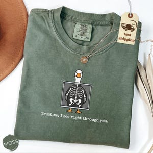 May include: A moss green Comfort Colors t-shirt featuring a cartoon duck x-ray design. The duck is inside a gray frame, with the text "Trust me, I see right through you." printed below. A gold necklace and a tag with "fast shipping" are also visible.