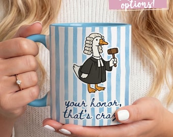 May include: A ceramic mug with a blue handle and rim, featuring a cartoon goose dressed as a judge holding a gavel. The mug has blue and white vertical stripes and the text "your honor, that's crazy".