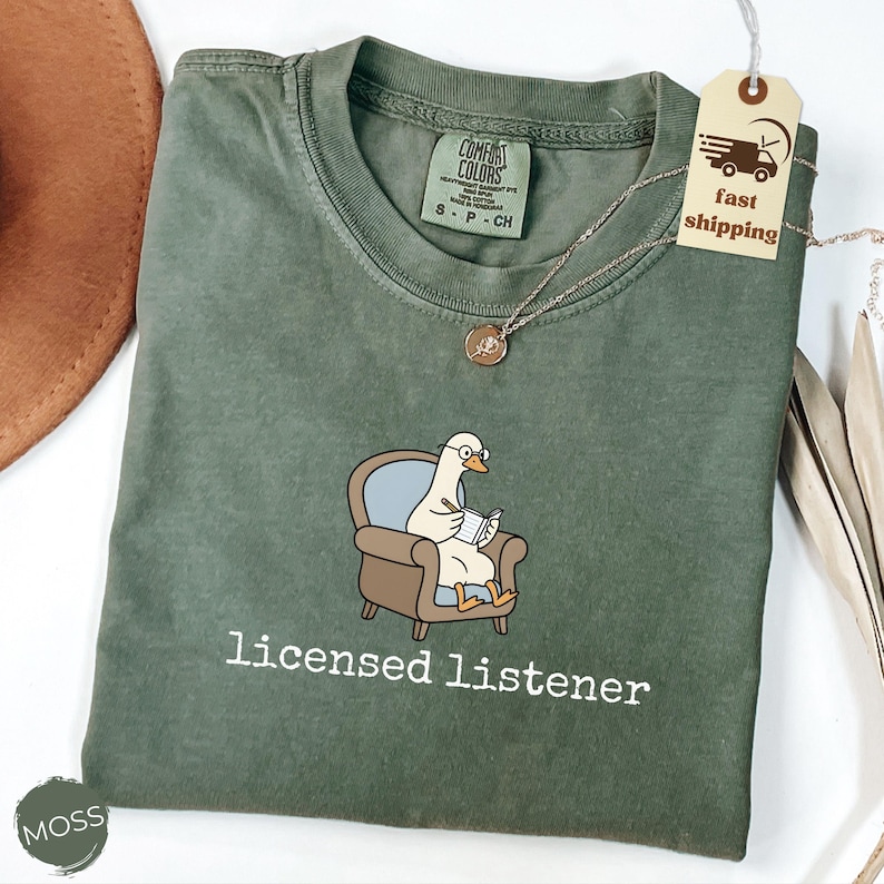 May include: Moss green Comfort Colors t-shirt featuring a cartoon duck wearing glasses and seated in a brown armchair, writing in a notepad. The text "licensed listener" is printed below the graphic. A gold necklace and a tag that says "fast shipping" are also visible.