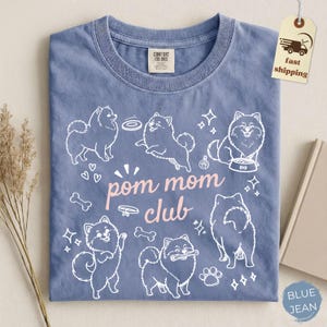 以下が含まれることがあります： クルーネックのジーンズブルーのTシャツ。Tシャツには、ポメラニアン犬、骨、足跡の白いグラフィックと、ピンク色の「pom mom club」の文字が描かれています。シャツにはComfort Colorsのタグが付いています。