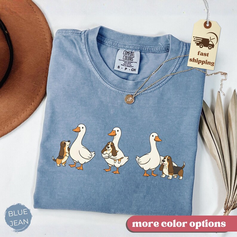 May include: Blue jean colored t-shirt with a crew neck. The shirt features a graphic of three white geese with orange beaks and feet, each accompanied by a brown and white dog. The shirt has the Comfort Colors brand label.