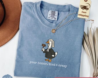 May include: Blue jean Comfort Colors t-shirt with a crew neck. The shirt features an illustration of a duck in a judge's robe holding a gavel, with the text "your honor, that's crazy". A gold necklace and a tag that says "fast shipping" are also visible.