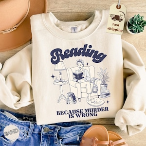 May include: Cream-colored sweatshirt with a navy blue graphic that reads "Reading" above an illustration of a person reading in a chair. The text "BECAUSE MURDER IS WRONG" is below the illustration. A fast shipping tag is visible.