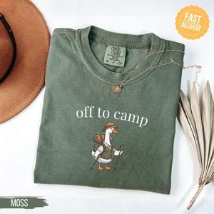 May include: Moss green t-shirt with the phrase "off to camp" in white. The shirt features a cartoon goose wearing a hat, backpack, and holding hiking sticks. A gold necklace is draped across the shirt.