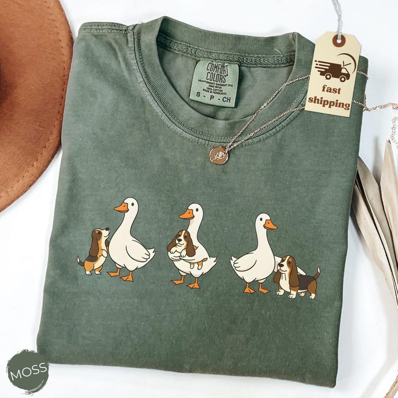 May include: Moss green Comfort Colors t-shirt featuring a graphic of three white geese with orange beaks and three basset hounds. The shirt has a crew neck and short sleeves. A silver necklace with a small pendant is also visible.