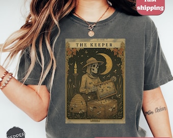 May include: Dark grey t-shirt featuring a tarot card design with the text "THE KEEPER". The card depicts a skeleton in a hat holding a honeycomb, with a crescent moon and bees. The shirt has a vintage, distressed look.