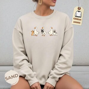 May include: Sand-colored sweatshirt featuring a whimsical design of three geese, each paired with a small dog. The dogs have different markings. The sweatshirt has the text "SAND" at the bottom left and a tag that says "Gildan 18000".