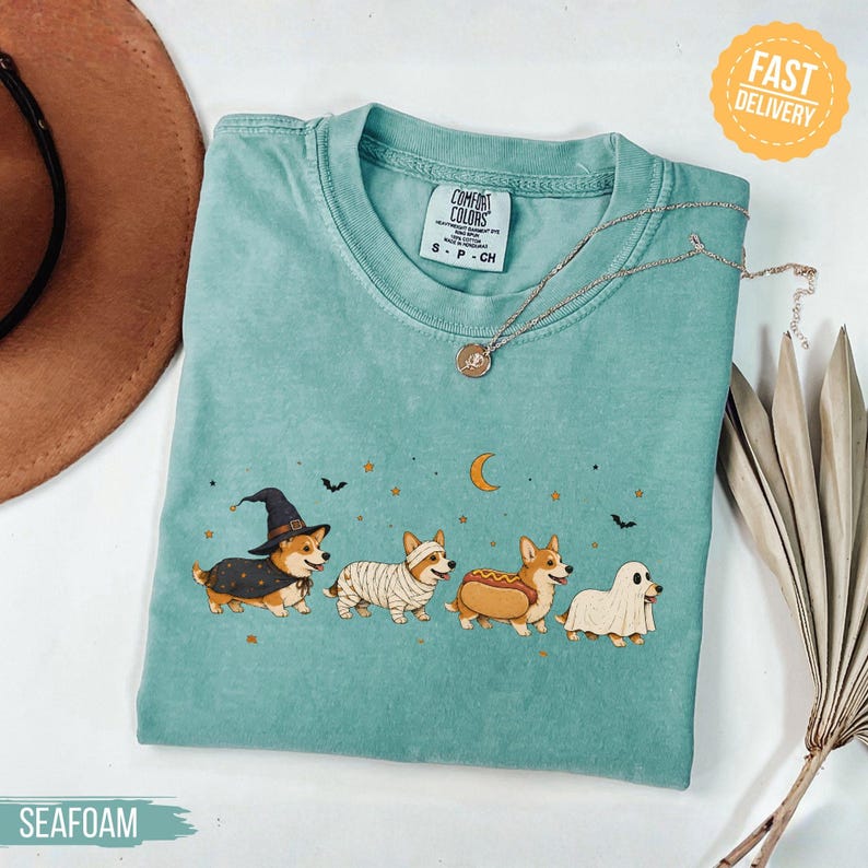 May include: A seafoam green t-shirt featuring a graphic of four corgis in Halloween costumes: a witch, a mummy, a hot dog, and a ghost. The design includes a crescent moon, stars, and bats. The shirt has a Comfort Colors label at the neck.