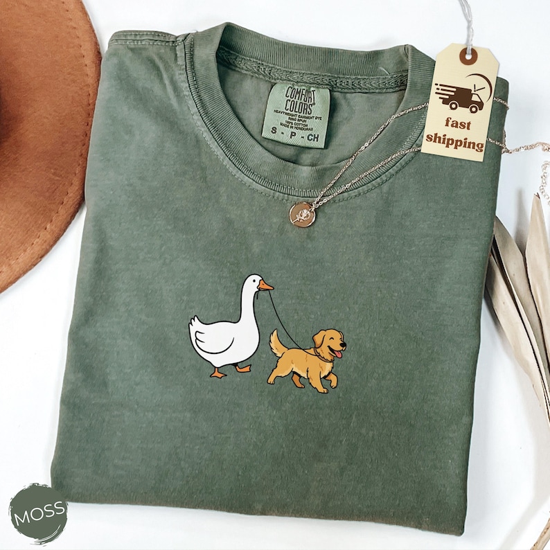 Pu&ograve; includere: Una maglietta Comfort Colors verde muschio con un disegno ricamato di un'oca bianca che porta a spasso un golden retriever al guinzaglio. La maglietta ha un girocollo e un'etichetta con la taglia. &Egrave; visibile anche una collana d'argento con un ciondolo.