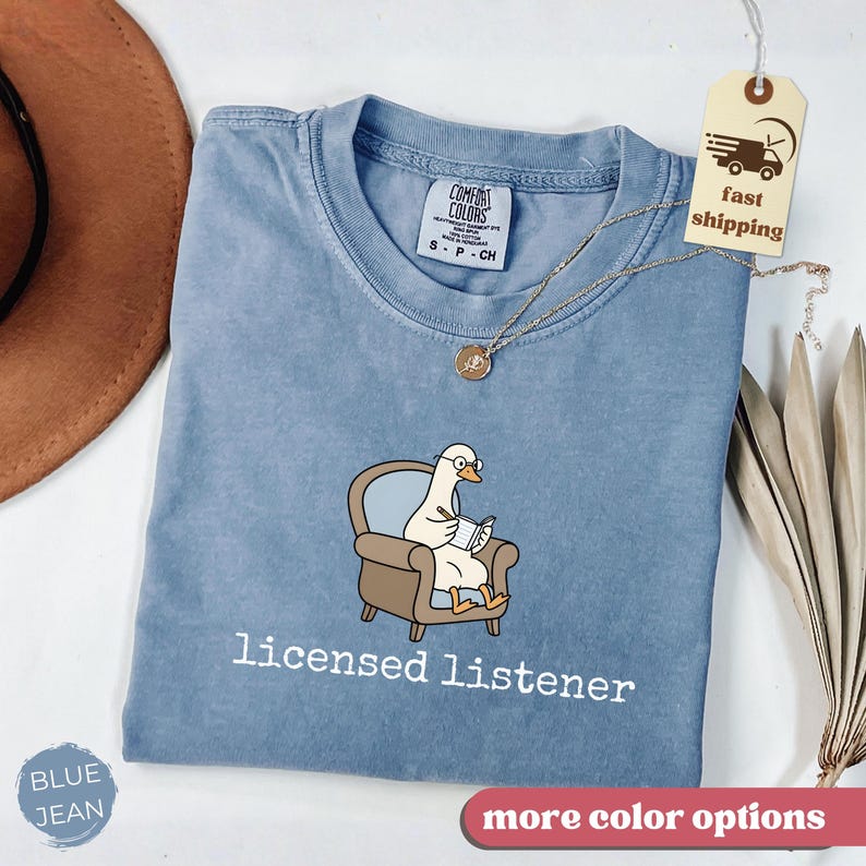 May include: A blue jean Comfort Colors t-shirt with a cartoon goose wearing glasses and reading in a chair. The text "licensed listener" is below the graphic. A gold necklace and a tag that says "fast shipping" are also visible.