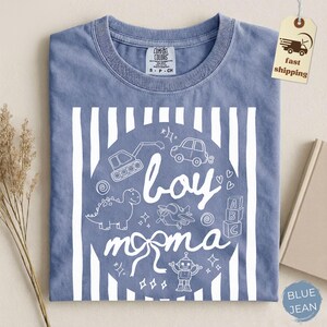 May include: Blue jean colored t-shirt with a white graphic. The graphic includes the words "boy momma" in a circle with drawings of a dinosaur, car, airplane, robot, and other toys. Vertical white stripes are on the shirt.
