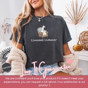 May include: Dark gray t-shirt with the text "licensed listener." The shirt features a cartoon goose wearing glasses and sitting in a chair, reading a book. The shirt is a comfortable, casual style.